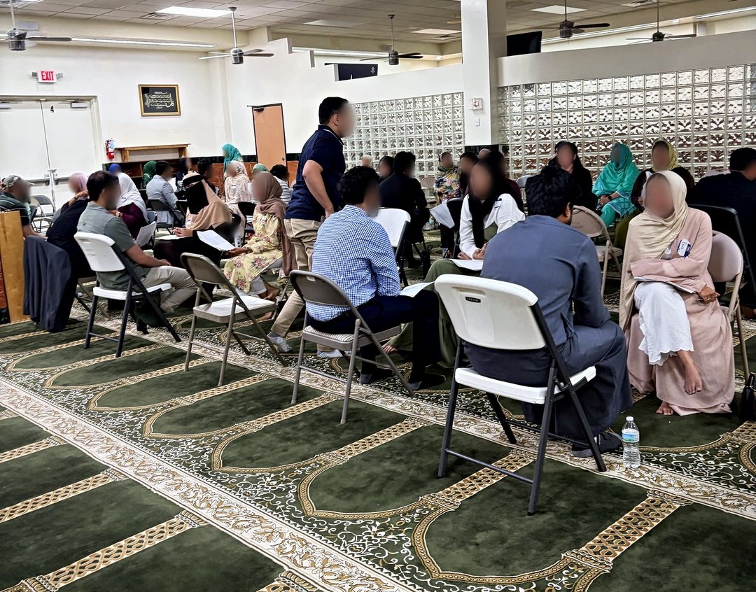 The far side of the hall at Masjid At-Taqwa, rows of candidates facing the glass block wall.